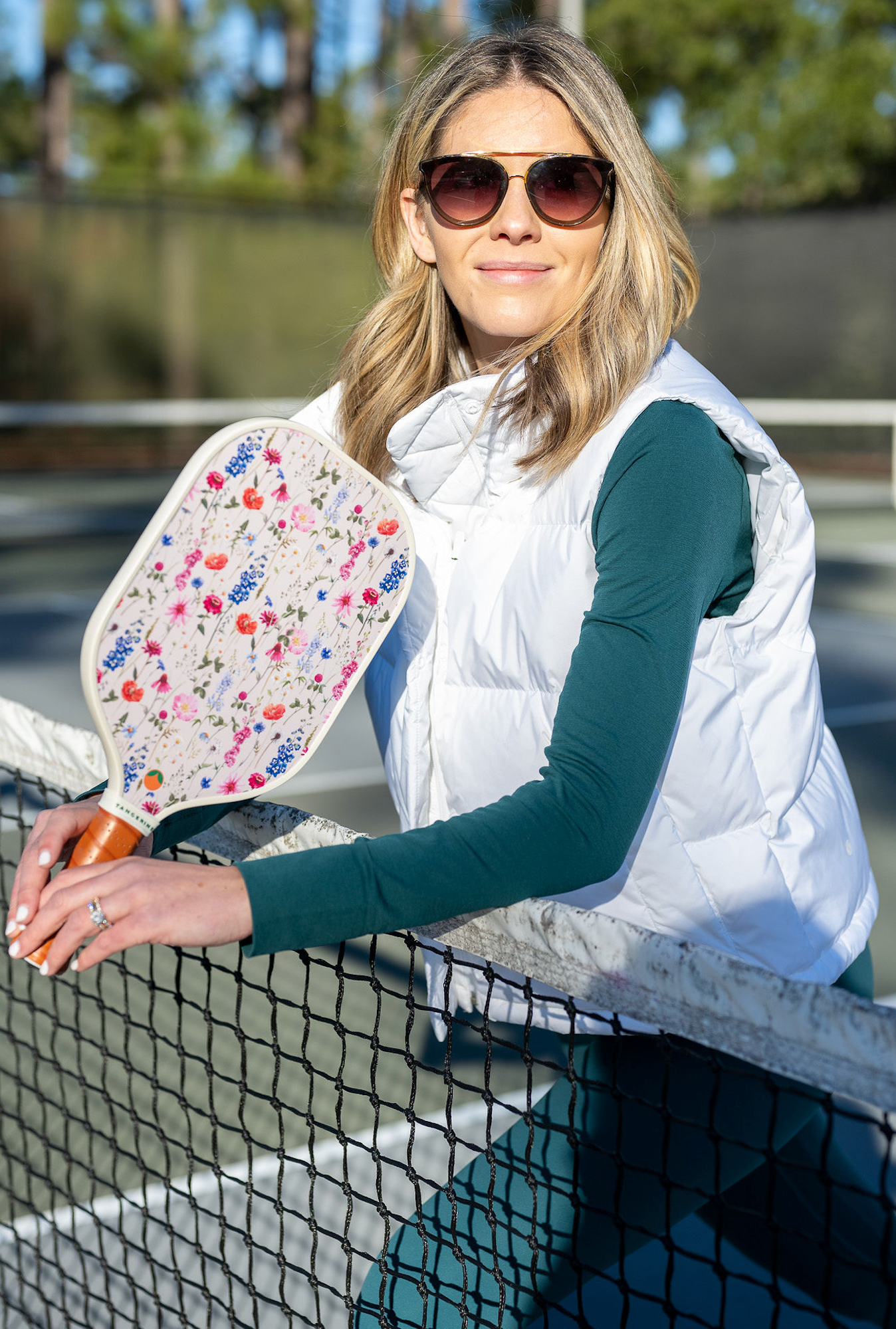 Woman in sunglasses posing on net with a floral pickleball paddle